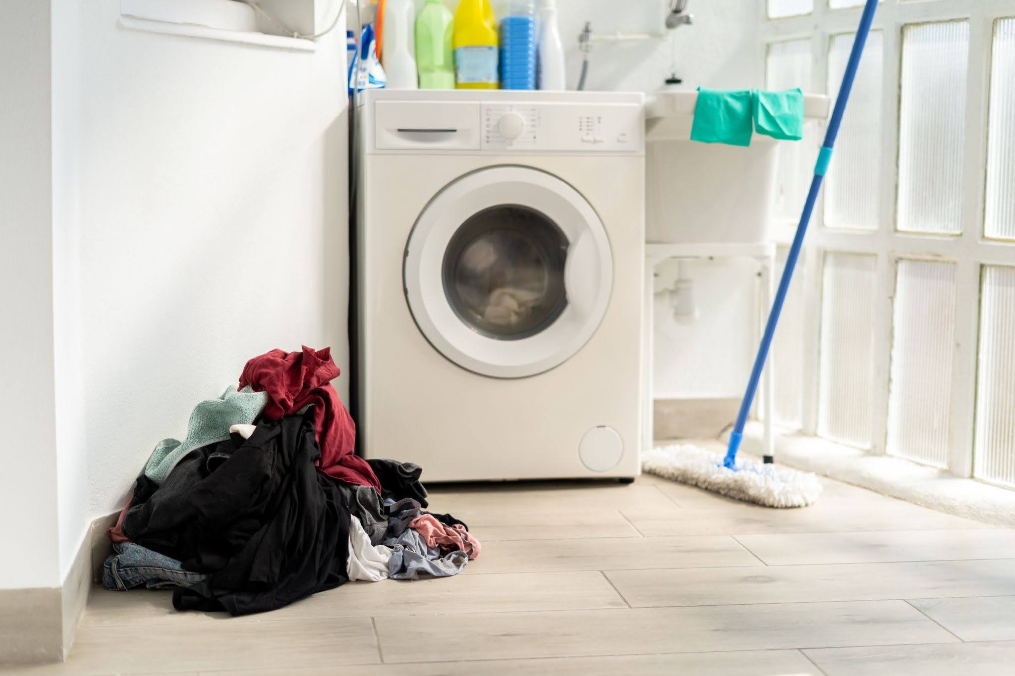 A pile of unwashed laundry building up beside a washing machine and mop in a home laundry room